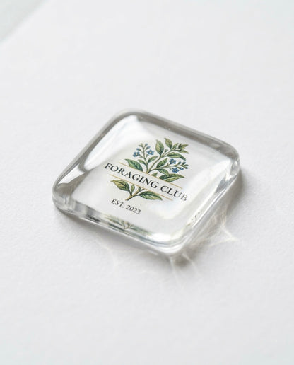 A single clear square resin sticker photographed at an angle on a white surface, featuring a botanical illustration of green leaves and small blue flowers above the text “Foraging Club Est. 2023” in elegant serif typography on a white background.